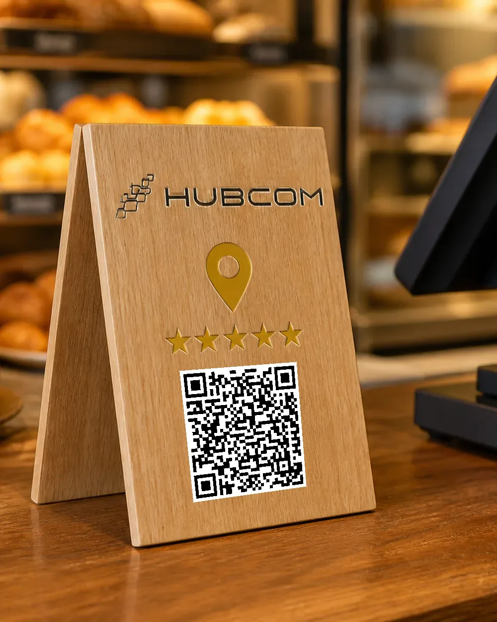 Standee gỗ sồi dựng trên quầy tiệm bánh cạnh máy tính tiền, mặt trước in logo Hubcom, ghim bản đồ vàng, năm ngôi sao và mã QR quét được dẫn tới trang đánh giá